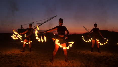 Un-Grupo-De-Hombres-Y-Mujeres-Muestran-Fuego-Por-La-Noche-En-La-Arena-Con-El-Telón-De-Fondo-De-Fuego-Y-Grúas-Torre.